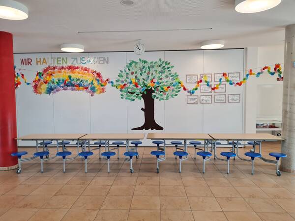 Aula mit Händen, die einen Regenbogen formen, einem Baum, Schulregeln und dem Spruch: "Wir halten zusammen!"