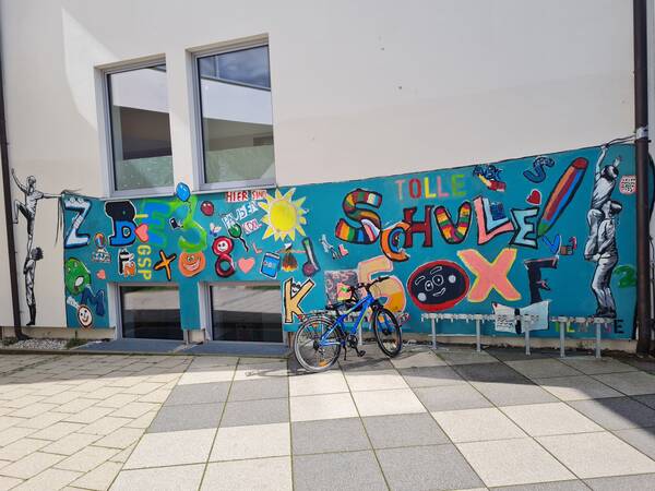 Graffiti im Innenhof mit Kindern, die ein Banner mit vielen Buchstaben und dem Spruch "Tolle Schule" aufhängen.