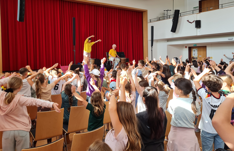 Übung im Bürgersaal mit Alma und Johnny auf der Bühne. Alma macht Bewegungen vor, die Kinder nachahmen.